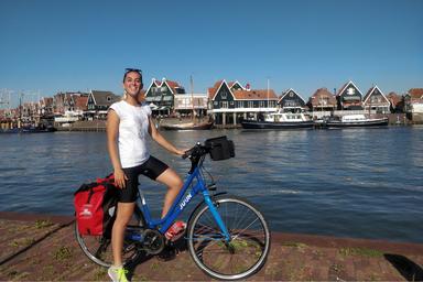 Bicicletta blu e borsa rossa, una ragazza con un volto sorridente e occhiali da sole sulla testa, si trovano su una banchina di una città di pescatori
