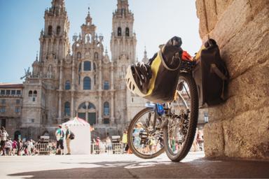 Bicicletta da corsa con borse laterli gialle e casco bianco appeso, è appoggiata ad un muro, sullo sfondo la celeberrima cattedrale di Santiago di Compostela