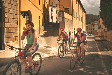 Quattro ciclisti stanno mangiando una pizza mentre pedalano lungo una delle strade della città di Buonconvento