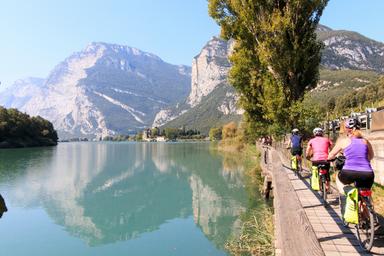 Specchio d'acqua sul quale si riflettono le montagne delle Dolomiti e un gruppo di ciclisti sono sulla pista ciclabile accanto al lago