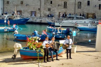 Un gruppo di musicisti in Puglia, stanno suonando una canzone tradizionale. Un'insenatura dove si sviluppa un piccolo porticciolo dove barche bianche e blu sono ormeggiate